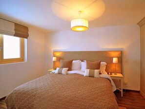 Quarto de hotel com uma cama grande, duas mesas de cabeceira, uma janela com cortinas e uma luminária no teto.