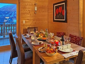 A wooden dining table set with plates, glasses, and candles, with a mountain view through a glass door. - Chalet Ourson Blanc by Interhome (Hérémence)