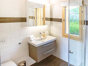 Ein modernes Badezimmer mit einer Duschkabine aus Glas, einem weißen Waschbecken mit Holzschrank und einem Fenster mit Blick auf Grünflächen.