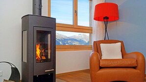 Ett rum med en vedeldad kamin, en fåtölj i läder, en röd golvlampa och ett fönster med utsikt över bergen.