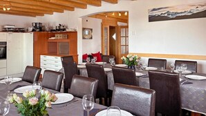 A dining area with tables set for four, wooden beams on the ceiling, a kitchen in the background, and a painting on the wall. - Chalet Julia by Interhome (La Tzoumaz)