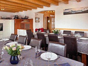 A dining area with tables set for four, wooden beams on the ceiling, a kitchen in the background, and a painting on the wall. - Chalet Julia by Interhome (La Tzoumaz)