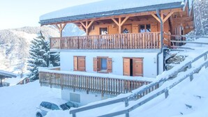 En tvåvånings chalet i trä med balkonger, träräcken och en snötäckt bergskedja i bakgrunden.
