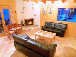 A cozy living room with a fireplace, leather sofa, wooden coffee table, and a large window with a view. - Chalet Maria by Interhome (La Tzoumaz)