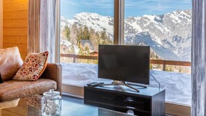 Ein Zimmer mit Fernseher auf einem Schrank, einer Couch mit einem gemusterten Kissen und Blick auf verschneite Berge durch große Fenster.
