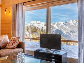 Ein Zimmer mit Fernseher auf einem Schrank, einer Couch mit einem gemusterten Kissen und Blick auf verschneite Berge durch große Fenster.