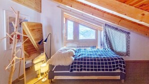 Ein Schlafzimmer mit schrägen Wänden, einem Bett mit karierter Bettdecke, einem Fenster mit Vorhängen und einer Lampe.