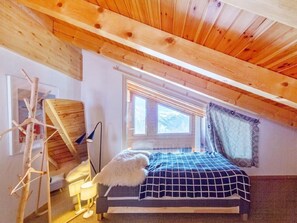 Ein Schlafzimmer mit schrägen Wänden, einem Bett mit karierter Bettdecke, einem Fenster mit Vorhängen und einer Lampe.