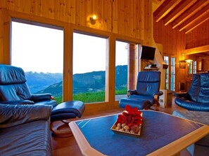 A room with large windows, leather chairs, a coffee table with a red flower arrangement, and a television on a wooden stand. - Chalet Alpina by Interhome (La Tzoumaz)