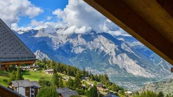 Blick auf Berge vom hölzernen Balkon, der über ein Tal mit Häusern und blauem Himmel blickt.