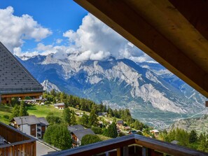 Blick auf Berge vom hölzernen Balkon, der über ein Tal mit Häusern und blauem Himmel blickt.