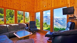 A wooden cabin interior with a sofa, armchairs, a TV, and large windows offering mountain views. - Chalet Alpina by Interhome (La Tzoumaz)