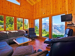 Ein Holzhütten-Interieur mit einer Couch, Sesseln, einem Fernseher und großen Fenstern mit Blick auf die Berge.