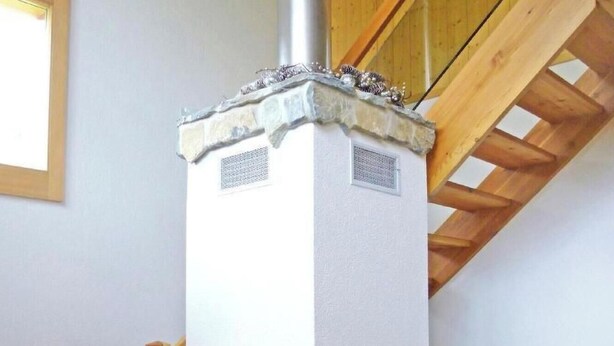A wood-burning stove with a stone base and a white upper section, located in a room with a wooden staircase and a black leather chair. - Chalet Chaud by Interhome (La Tzoumaz)