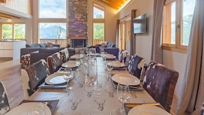 A dining area with a long table set for a meal, a stone fireplace, and large windows offering mountain views. - Chalet Harmonie by Interhome (La Tzoumaz)