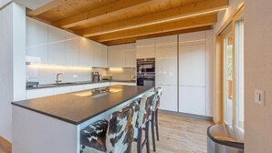 A modern kitchen with a central island, wooden ceiling beams, and a view of the outdoors. - Chalet Harmonie by Interhome (La Tzoumaz)