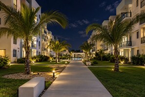 A nighttime view of a modern hotel with a central pool, surrounded by palm trees and well-lit pathways. - Your  Peaceful tropical  Punta Cana Getaway 2BR Condo with all amenities & more (Punta Cana)