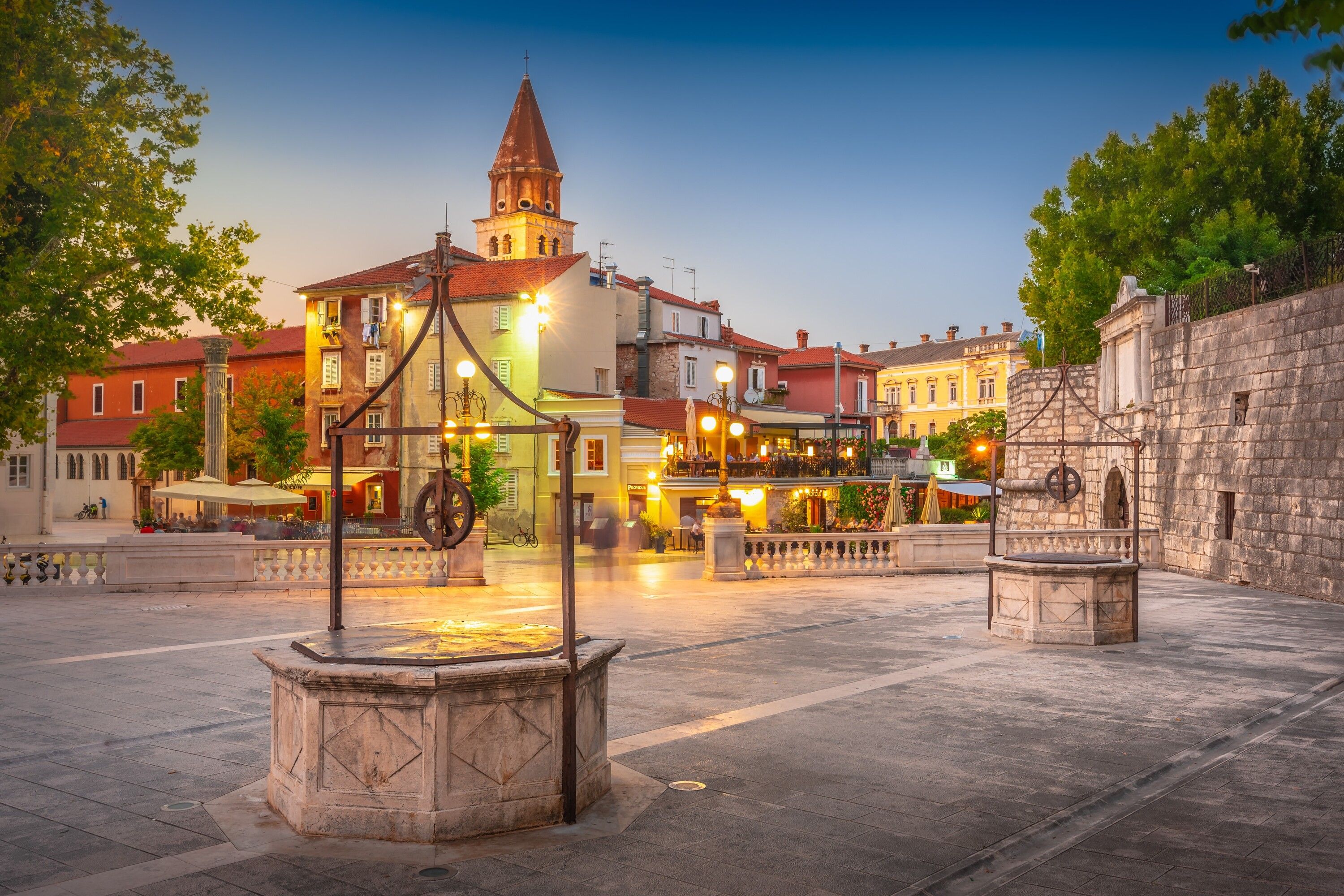 A historic square with a central fountain, a church with a bell tower, and surrounding buildings with balconies and outdoor seating.