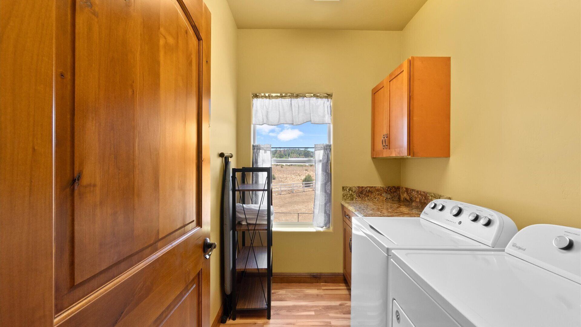 A laundry room with a washer and dryer, wooden cabinets, a window with curtains, and a shelf with a towel.