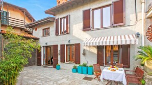 Une cour dallée de pierres, une table avec des chaises, des plantes en pot et un bâtiment avec des volets.