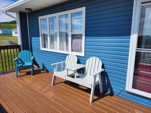 A blue house with a wooden deck, two white Adirondack chairs, and a small table.