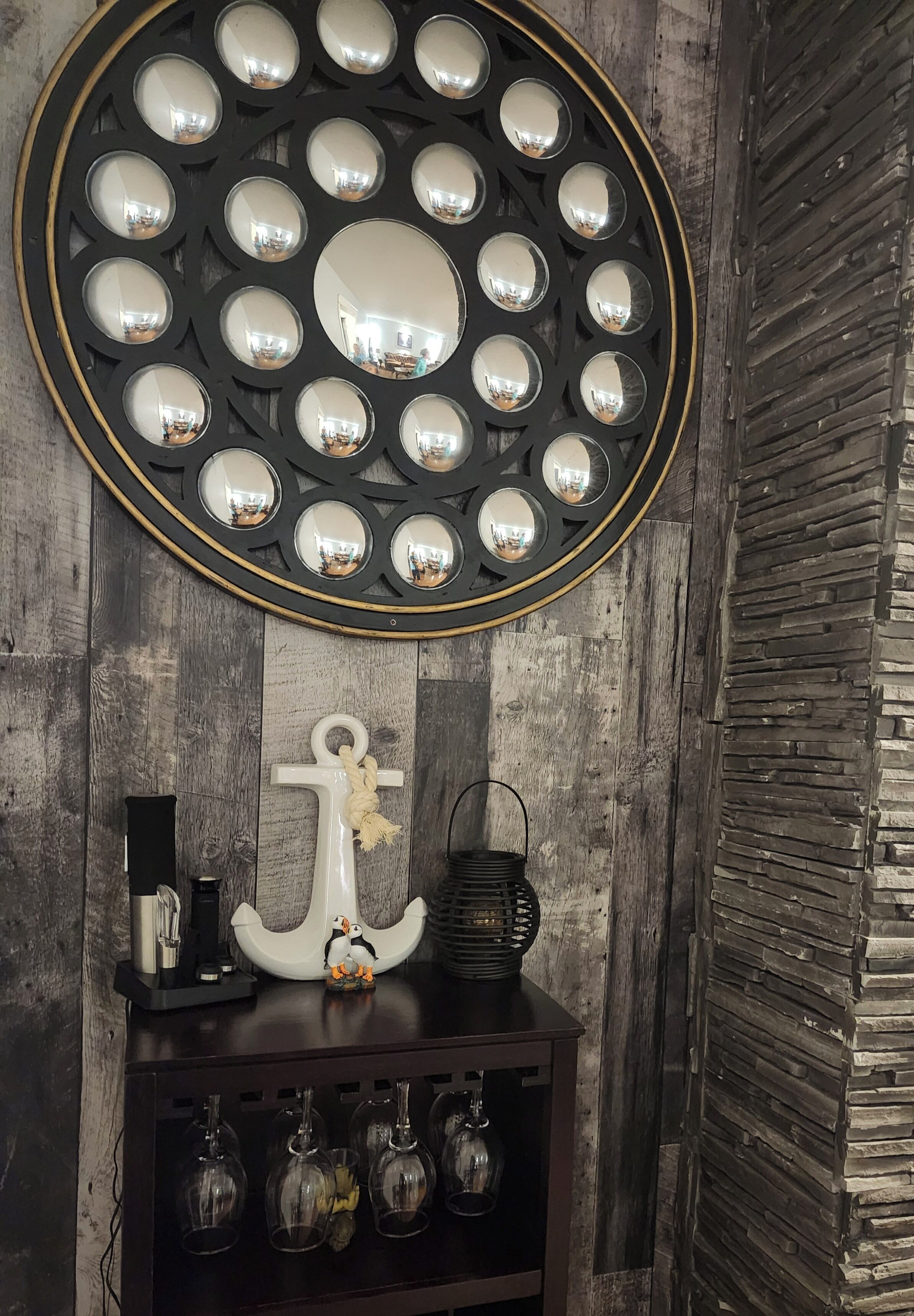 Un miroir mural avec un cadre circulaire, un mur en bois avec un motif texturé, une petite décoration en forme d’ancre blanche, une lanterne noire et un porte-bouteilles à vin avec des verres vides.