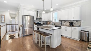 A modern kitchen with white cabinets, stainless steel appliances, a central island with stools, and a wooden staircase. - Luxury 6BR 3 BA Beach Home Sleeps 14 Huge Front Porch Walk To Beach and Downtown (Ocean City)