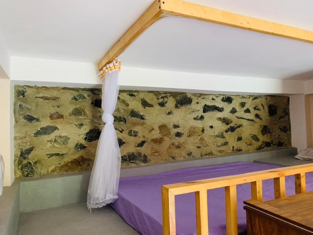 A room with a stone wall, a bed with a purple bedspread, a wooden bench, and a white canopy.