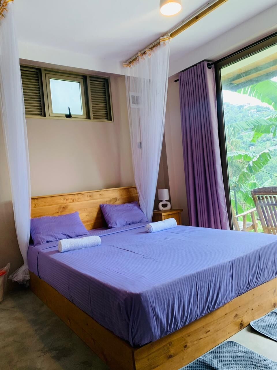 Een bed met houten frame en paars beddegoed, een nachtkastje met lamp, en een raam met uitzicht op groen.