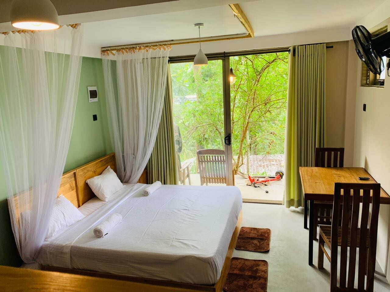 Une chambre avec un lit, une table, des chaises, un ventilateur et une vue sur la verdure.