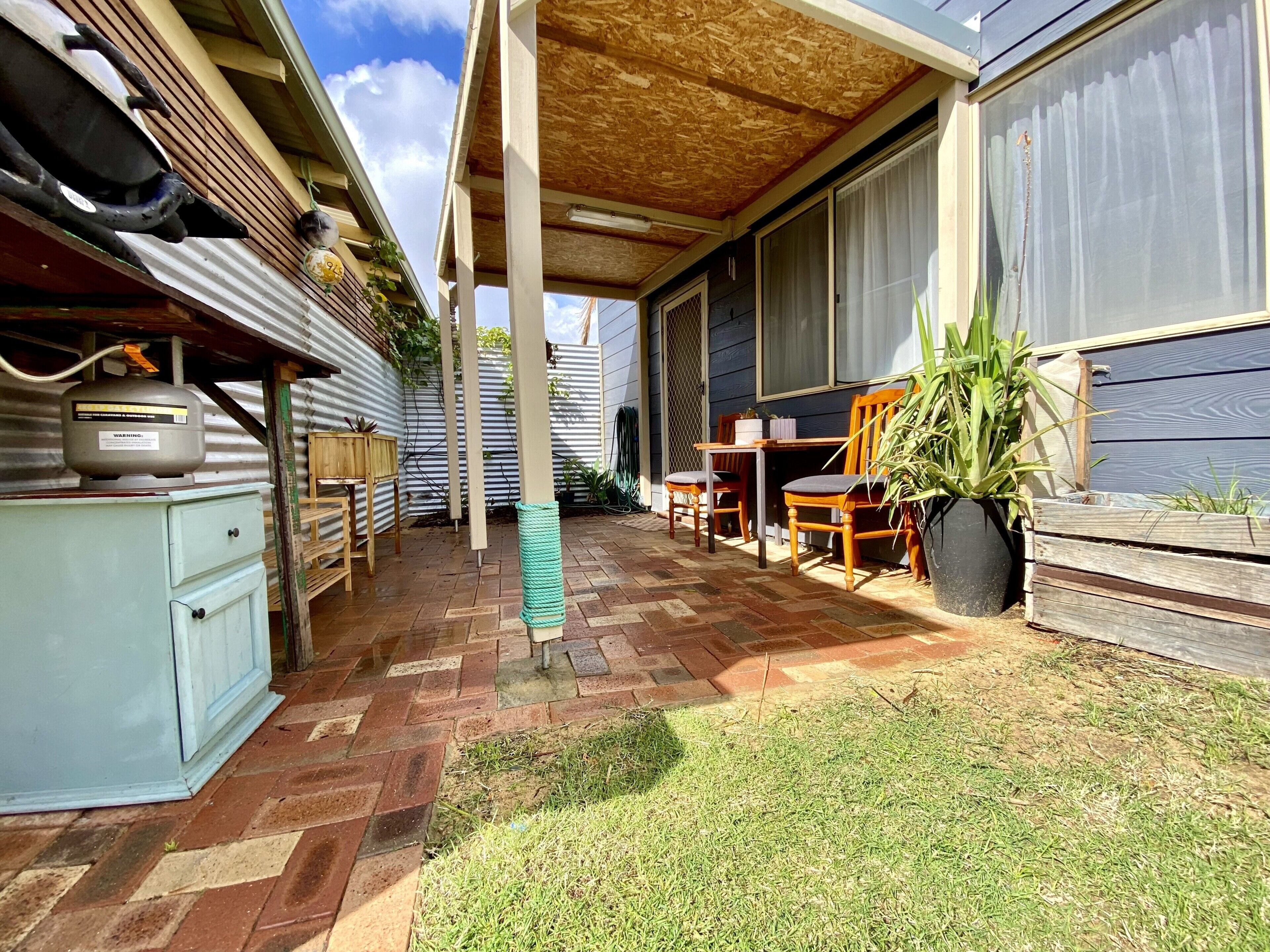 Ein überdachter Patio-Bereich mit Ziegelboden, Holzmöbeln und einem Blumentopf.