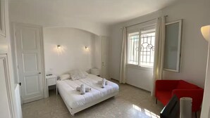 Ein Schlafzimmer mit einem Bett, Nachttischen, einem roten Sessel und einem Fenster mit Vorhängen.