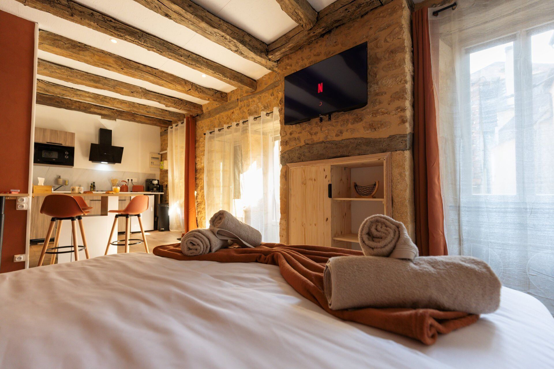 Une chambre d’hôtel avec un lit, un bureau, une chaise, un téléviseur fixé au mur et un coin cuisine à l’arrière-plan.