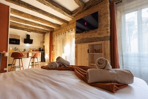 A hotel room with a bed, a desk, a chair, a TV mounted on the wall, and a kitchen area in the background. - Hostcare - Le Secret Sarladais – Downtown (Sarlat-la-Canéda)