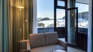 Ett hotellrum med en soffa, ett litet bord, ett element och stora fönster med utsikt över ett snötäckt landskap.