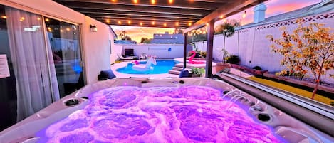 Un jacuzzi con un flotador de unicornio, una piscina y un patio con luces colgantes.