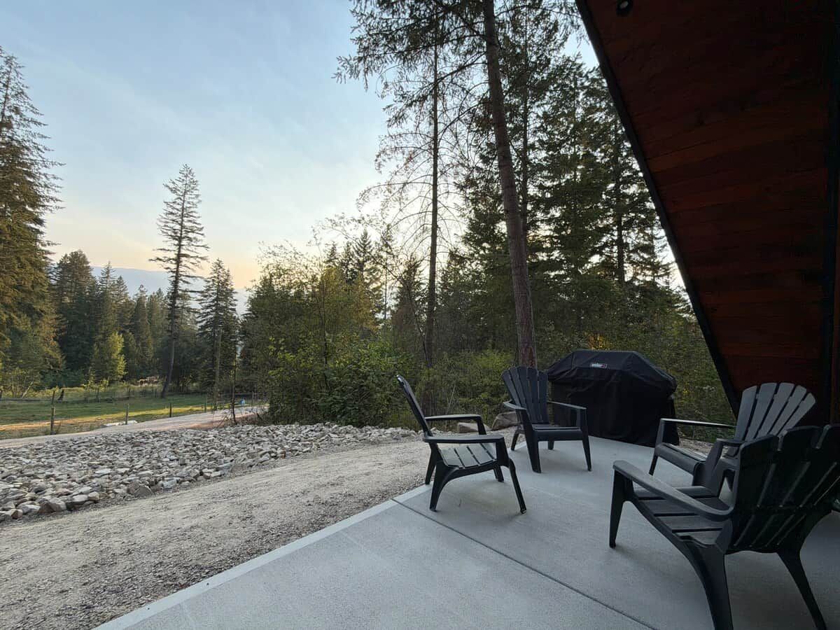 En udendørs terrasse med Adirondack-stole og grill, omgivet af et grusareal og en tæt skov.