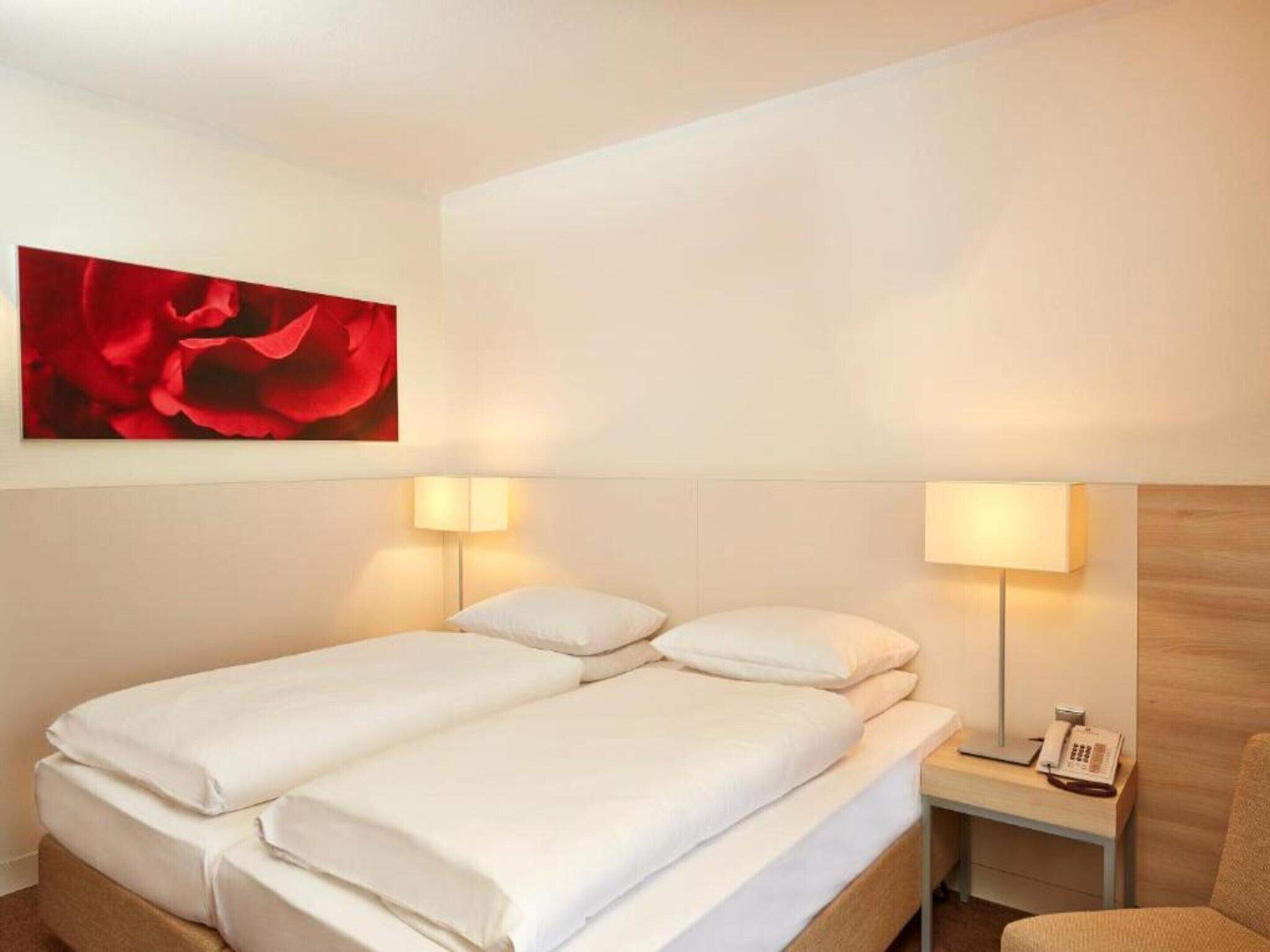 A hotel room with a double bed, two bedside lamps, a wooden nightstand with a phone, and a red flower painting on the wall.