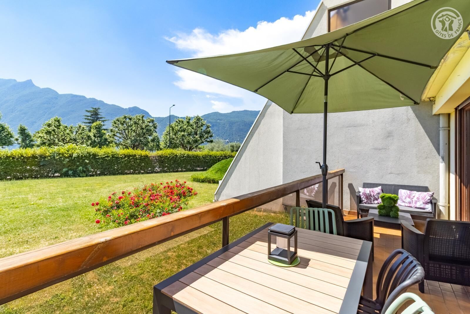 A balcony with a table, chairs, and an umbrella, overlooking a garden with flowers and a mountain view.