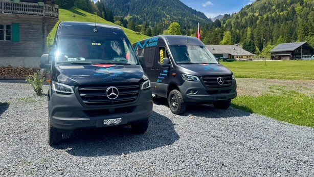 Duas carrinhas Mercedes estacionadas sobre um leito de cascalho, com montanhas ao fundo.