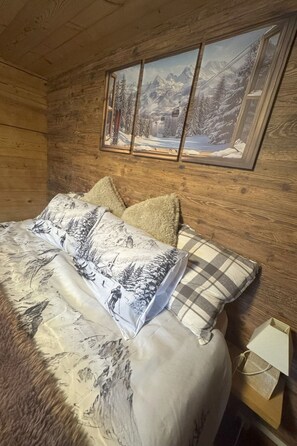 Ein Zimmer in einer Holzhütte mit einem Bett, Kissen, einem Gemälde einer verschneiten Berglandschaft und einer Lampe auf einem Nachttisch.
