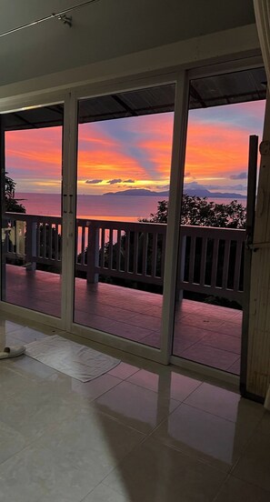 Habitación con vista a la puesta de sol sobre el océano, un balcón con barandilla y una puerta que da al exterior.