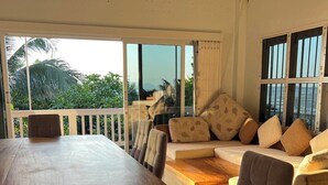 A room with a large wooden table, a sofa with patterned cushions, and a view of greenery and water through large windows.