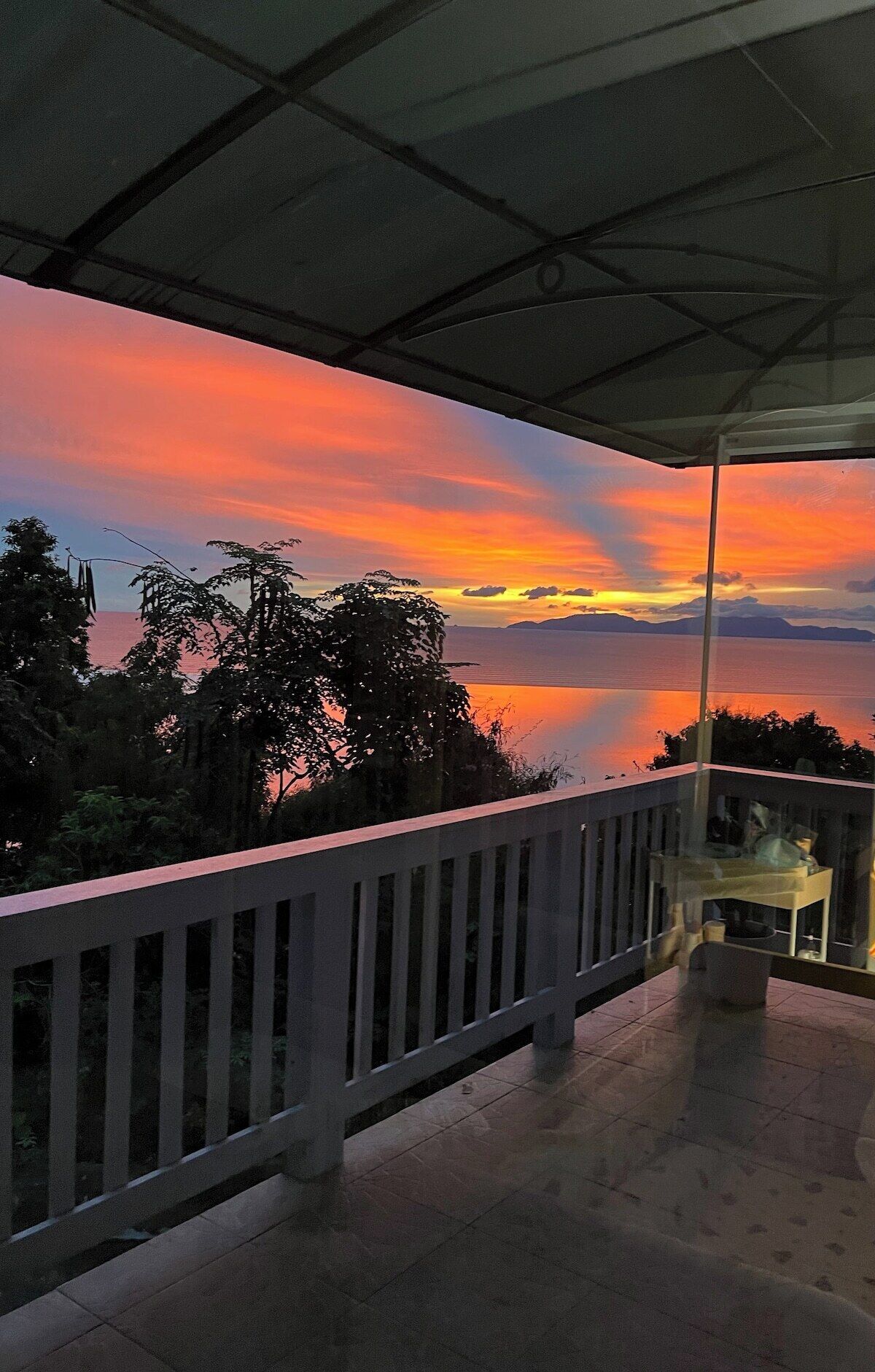 A balcony with a view of a sunset over a body of water, surrounded by trees.
