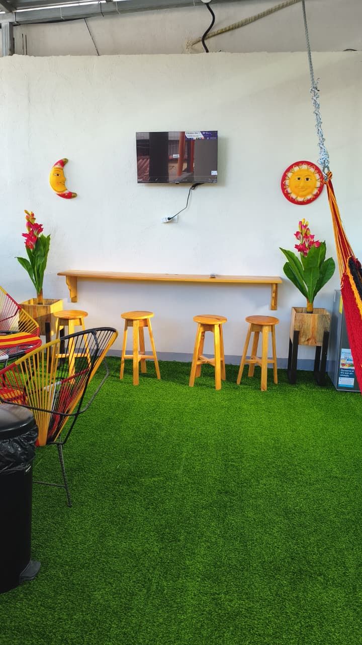 Ein Zimmer mit grünem Kunstgrase, einer hölzernen Bank, vier Holzstühlen, einem an der Wand montierten Fernseher und zwei Blumentöpfen mit roten Blüten.