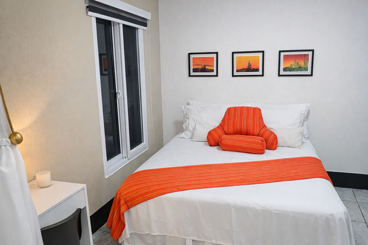 Un dormitorio con cama, ventana, mesita de noche y tres cuadros en la pared.