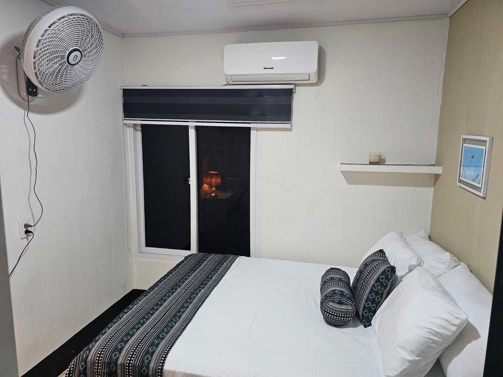 A bedroom with a bed, a standing fan, an air conditioner, a window with a blind, and a small shelf with a picture.