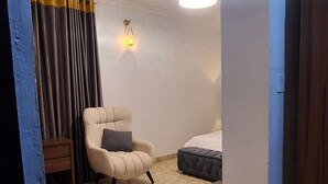 Ett rum med en beige fåtölj, en liten pall, ett nattduksbord, en lampa och en säng med en vit sänggavel.