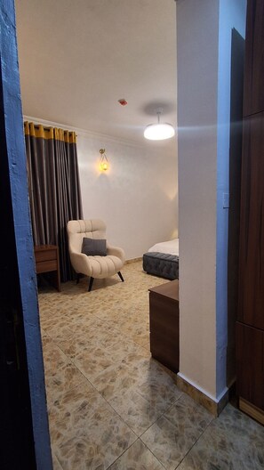 A room with a beige armchair, a small ottoman, a nightstand, a lamp, and a bed with a white headboard. - Lovely Spacious 3-bedroom apartment with AC in awesome Lagos, 15min from Airport (Lagos)
