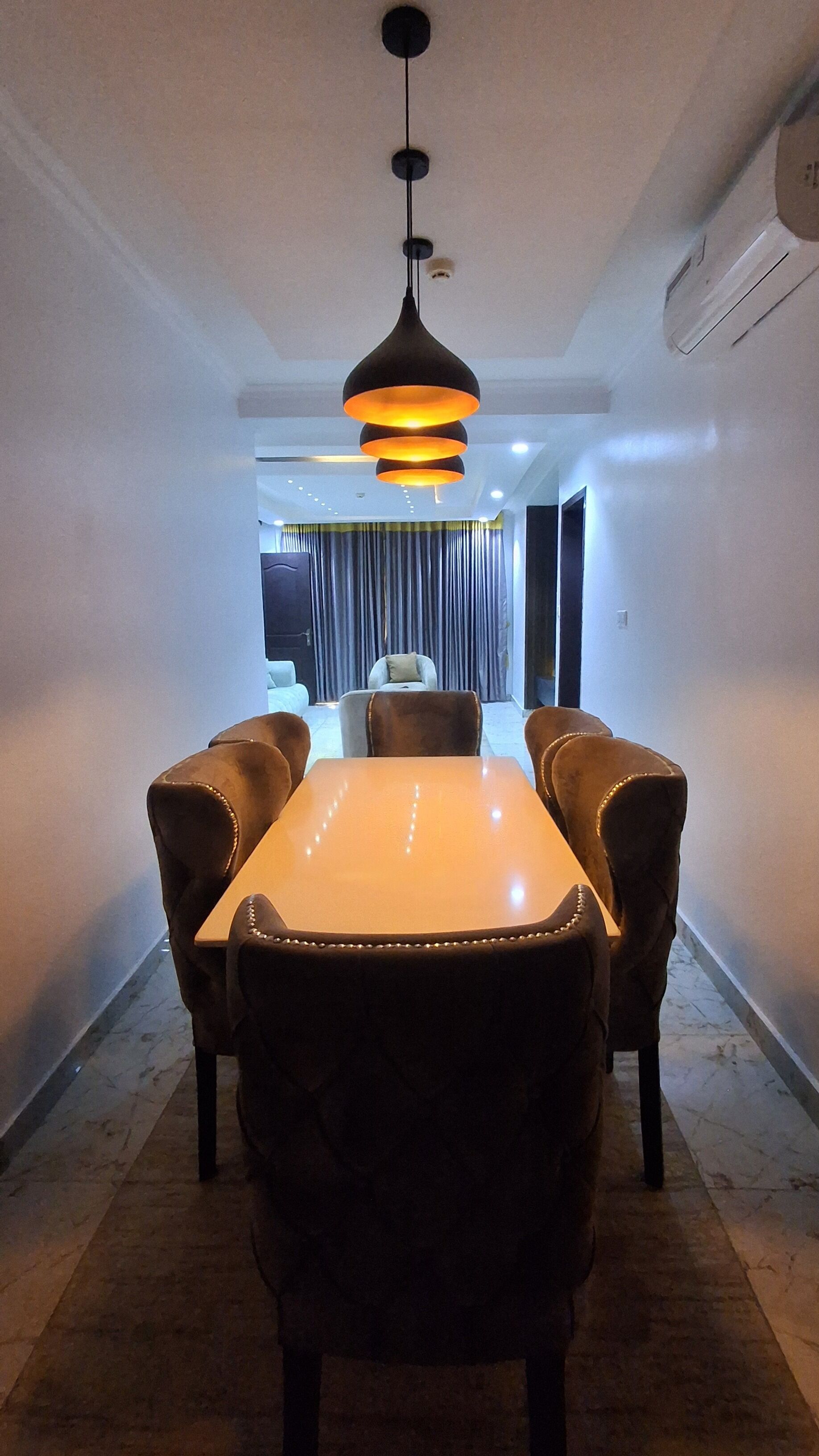 A dining area with a long table and upholstered chairs, a pendant light, and a view to a balcony with curtains.